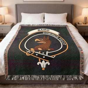 May include: A woven throw blanket with a dark green and purple plaid pattern. The blanket features a central emblem with a brown eagle, a white banner with the name "BAIRD", and the Latin phrase "DOMINUS FECIT". The edges have a fringed trim.