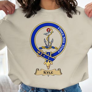 May include: Beige sweatshirt featuring a blue and gold crest with an anchor and the words "Fides Non Timet." Below the crest is a banner with the name "Kyle." The sweatshirt is a long-sleeved crew neck.