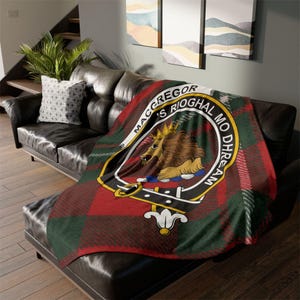 May include: A red, green, and black plaid throw blanket draped over a black leather couch. The blanket features a crest with a lion and the text "MACGREGOR'S RIOGHAL MO DHREAM". The blanket is a cozy home decor item.