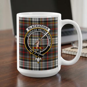 May include: A white ceramic mug featuring a grey, black, red, and yellow tartan plaid pattern. Centered on the mug is a circular crest with a black belt design, the text BLACKADDER COURAGE HELPS FORTUNE, and an illustration of a hand holding a sword.