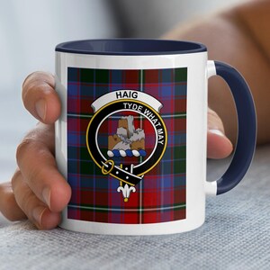 May include: White ceramic mug with a navy blue handle and rim. The mug features a colorful plaid pattern and a crest with the words "HAIG" and "TYDE WHAT MAY". The mug is held by a person.