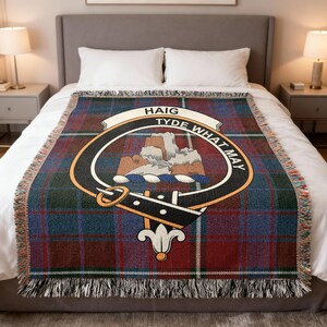 May include: A woven throw blanket with a plaid pattern in shades of red, blue, and green. The blanket features a central emblem with the text "HAIG TYDE WHAT MAY". The blanket has a fringed edge and is displayed on a bed.