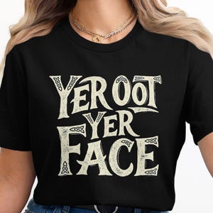 May include: Black t-shirt featuring the phrase "YER OOT YER FACE" in a distressed, off-white font. The text is in three lines with Celtic knot designs. The shirt has a crew neck.
