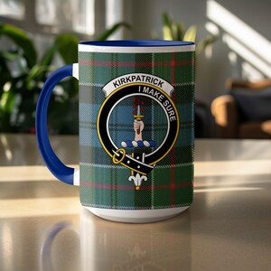 May include: White ceramic mug with a blue handle and rim. The mug features a green, blue, and red plaid pattern with a crest that reads "Kirkpatrick I Make Sure". The crest includes a hand holding a sword.