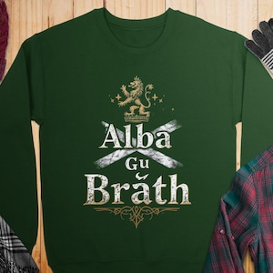 Puede incluir: Una sudadera verde con un diseño blanco y dorado que presenta un león, una cruz y el texto "Alba Gu Brath".