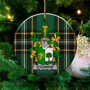 May include: A round ceramic ornament featuring a green and black plaid tartan pattern. Centered is a heraldic coat of arms with a silver helmet, green and gold mantling, and a shield displaying a lion and a tree. The name O'CONNOR is printed below.