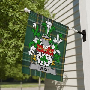 May include: A green and yellow plaid flag with a family crest. The crest features a helmet, red and white shield, and the name "LEECH" on a banner. The flag is mounted on a white pole and black bracket.