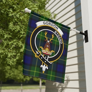 May include: A rectangular flag with a green, blue, and black tartan pattern. The flag features a circular emblem with a deer and the words "GORDON BYDAND". The flag is mounted on a white pole attached to a white wall.
