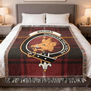 May include: A tartan throw blanket in red, black, and yellow, with a fringed border. It displays a circular emblem with a boar and the words "MACIVER NUNQUAM OBLIVISCAR". The blanket is placed on a bed.