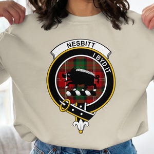 May include: Beige sweatshirt featuring a circular emblem with the name "NESBITT" above and "I BYD IT" below. The emblem includes a black boar, a red and green plaid pattern, and a gold and black border.