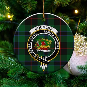 May include: Circular Christmas decoration with a green, black, and red tartan design. It displays a crest with the name "DOUGLAS" and the motto "NEVER BEHIND JAMAIS ARRIERE". The ornament is suspended on a Christmas tree.