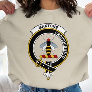 May include: Beige sweatshirt featuring a circular emblem with the name "MAXTONE" above a bee illustration. The emblem includes the words "PROVIDUS ESTO" and is surrounded by a black and gold border. The bee has yellow and black stripes.