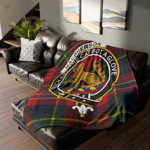 May include: A tartan throw blanket in red, green, and yellow, draped over a black leather sofa. The blanket displays a crest with a golden cat and the words "Macpherson The Cat Bot a Glove."