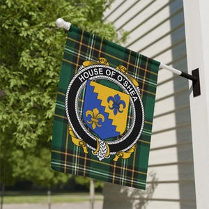 May include: A rectangular flag featuring a green, black, and yellow tartan pattern. Centered on the flag is a circular emblem with the text HOUSE OF O'SHEA, enclosing a shield divided diagonally with blue and yellow sections and two fleur-de-lis.