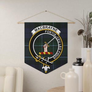 Puede incluir: Un tapiz de pared con el escudo de armas de la familia MacBrayne sobre un fondo de cuadros verdes, azules y negros. El escudo incluye una espada, una mano y las palabras "MACBRAYNE FORTIS CEU LEO FIDUS" en un diseño circular. La pancarta está suspendida de una barra de madera.