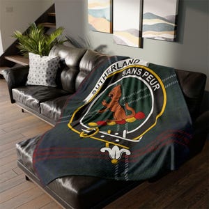 May include: A green and red tartan blanket with the Sutherland family crest, featuring a brown animal and the words "SUTHERLAND SANS PEUR" in a white circle. The blanket is on a black leather sofa.