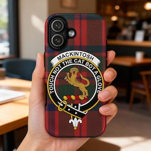 May include: A phone case with a red, green, and blue plaid pattern. The case features a crest with a lion and the words "MACKINTOSH TOUCH NOT THE CAT BOT A GLOVE". The phone case is held in a person's hand.