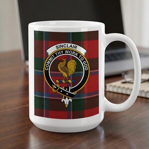 May include: A white ceramic mug featuring a Sinclair clan crest with a golden rooster emblem inside a black belt design. The text reads SINCLAIR and COMMIT THY WORK TO GOD. The background of the emblem is a red, green, and blue tartan plaid pattern.