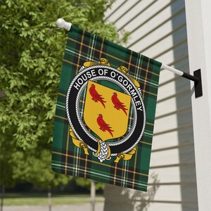 May include: A rectangular flag featuring a green, black, and yellow tartan pattern. Centered is a circular black belt with the text HOUSE OF O'GORMLEY in white, enclosing a yellow shield displaying three red birds arranged in a triangular formation.