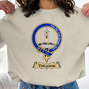 May include: Cream-colored sweatshirt featuring a blue and gold crest design. The crest includes a hand holding a sword, surrounded by text. The banner below reads "Treanor".