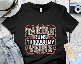 May include: Black t-shirt with the text "Tartan Runs Through My Veins" in a decorative design. The text features a plaid pattern in red, green, and white. The shirt is displayed with a hat, sunglasses, and jeans.