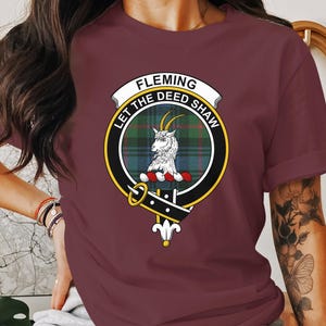 May include: Burgundy t-shirt with a Fleming family crest design. The crest features a goat, a tartan pattern, and the words "FLEMING", "LET THE DEED SHAW". The crest is framed by a gold and black border.