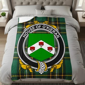 Puede incluir: Manta a cuadros verdes con las palabras "HOUSE OF O'KIERAN" que rodean un escudo con un fondo verde y tres figuras rojas con forma de gato. La manta tiene un borde blanco en forma de cuerda y detalles dorados.