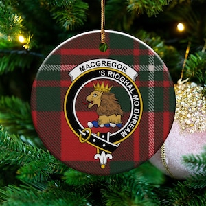 May include: Round Christmas ornament with a red, green, and white plaid pattern. The ornament features a crest with a lion and the words "Macgregor's Rioghal Mo Dhream". A gold string is attached for hanging. The ornament is displayed on a Christmas tree.