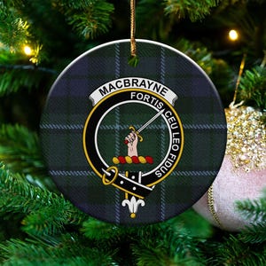 Puede incluir: Adorno navideño redondo con el escudo de la familia MacBrayne, sobre un fondo de cuadros verdes y azules. El escudo incluye una mano sosteniendo una espada, con las palabras "MACBRAYNE FORTIS CEU LEO FIDUS". El adorno cuelga de un hilo dorado.