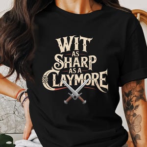 May include: Black t-shirt with the text "Wit as Sharp as a Claymore" in a vintage style font. The design includes two crossed swords. The shirt is a classic crew neck.