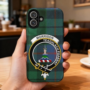 May include: A green plaid phone case featuring the Morrison clan crest. The crest is circular with a sword, a crown, and the words "MORRISON TEAGHLACH PHABBAY". The case has a camera cutout and is held in a hand.
