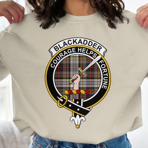May include: Cream-colored sweatshirt featuring a circular emblem with the words "BLACKADDER COURAGE HELPS FORTUNE." The emblem includes a tartan pattern, a hand holding a sword, and a decorative border. The sweatshirt is a casual garment.
