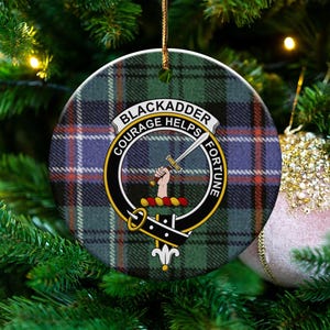 May include: Round Christmas ornament with a tartan pattern in green, blue, and red. The ornament features the text "BLACKADDER COURAGE HELPS FORTUNE" around a crest with a sword and hand. The ornament hangs from a gold string.