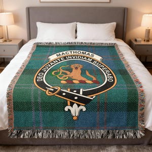 May include: A green and blue tartan throw blanket with a fringed edge. The blanket features a crest with the text "MACTHOMAS" and a lion emblem. The blanket is displayed on a bed with white bedding.