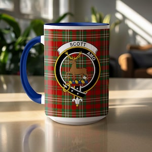 May include: A white ceramic mug with a blue handle and rim. The mug features a red and green plaid pattern with a crest that reads "SCOTT AMO" and a brown deer illustration. The mug is sitting on a light-colored surface.