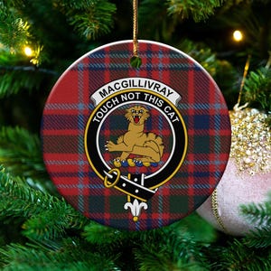 Puede incluir: Adorno navideño redondo con un patrón de cuadros rojos, azules y verdes. El centro presenta un escudo con un león y el texto "MACGILLIVRAY TOUCH NOT THIS CAT". El adorno cuelga de una cuerda dorada.