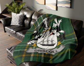 Könnte beinhalten: Eine grün-gelb karierte Decke mit dem Driscoll-Familienwappen, drapiert über einem schwarzen Ledersofa. Das Wappen zeigt ein Schiff, einen Schild und den Namen "DRISCOLL". Die Decke hat ein weiches, strukturiertes Aussehen.