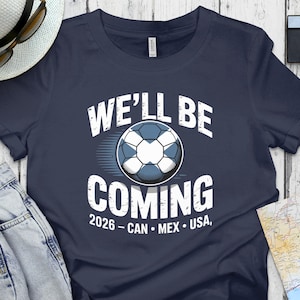 May include: Navy blue t-shirt with the text "WE'LL BE COMING" above a soccer ball graphic. Below the graphic is the text "2026 - CAN • MEX • USA". The shirt is next to a pair of blue jeans, a hat, sunglasses, a camera, and a map.