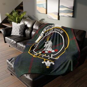 May include: A tartan blanket in green, navy, and red, with a white and gold emblem of a goat. The blanket is draped over a black leather sofa. The emblem reads "FLEMING THE DEEP SHAW".