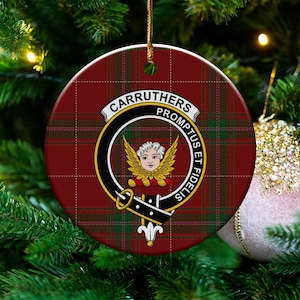 May include: Round Christmas ornament with a red and green plaid pattern. The ornament features a crest with the words "CARRUTHERS PROMPTUS ET FIDELIS" and a winged figure. A gold string is attached for hanging.