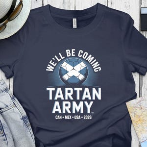 May include: Dark blue t-shirt featuring the text "WE'LL BE COMING" above a football with a Scottish flag design. Below the ball, the text reads "TARTAN ARMY" and "CAN • MEX • USA • 2026".