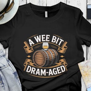 May include: Black t-shirt with a graphic design featuring a whiskey barrel, a glass of whiskey, and the text "A Wee Bit Dram-Aged" in a vintage style. The design is in shades of brown, beige, and white.