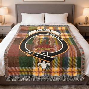 May include: A woven throw blanket with a plaid pattern in shades of brown, green, and yellow. The center features a crest with the words "Macgill" and "Sine Fine". The blanket has a fringed edge and is displayed on a bed.