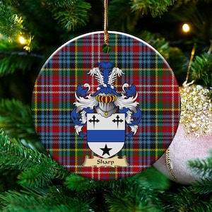 May include: A round ceramic ornament featuring a red, blue, green, and yellow tartan plaid pattern. Centered is a coat of arms with a silver helmet, blue and white mantling, and a shield displaying two black crosses, a blue bar, and a black star.