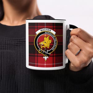 May include: A white ceramic mug with a black handle and rim, featuring a red and black tartan pattern. The mug displays a circular emblem with a yellow boar and the words "MACIVER NUNQUAM OBLIVISCAR".