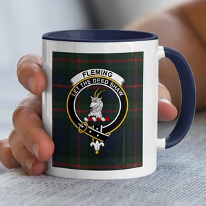 May include: White ceramic mug with a navy blue handle and rim. The mug features a tartan pattern with a crest that reads "FLEMING LET THE DEED SHAW". The crest includes a goat and a banner.