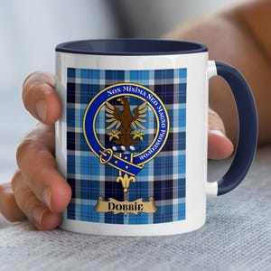 May include: White ceramic mug with a blue handle and rim, featuring a blue and white plaid pattern. The mug displays a circular emblem with a brown eagle and the name "Dobbic" on a banner.