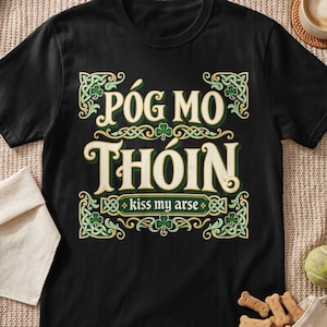 May include: A black short-sleeved t-shirt featuring a graphic print with the text PÓG MO THÓIN and kiss my arse in a cream-colored font. The design is surrounded by an intricate green and gold Celtic knotwork border with small green shamrock icons.
