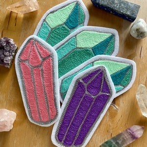 Pode incluir: Quatro patches de feltro bordados em forma de cristais. Os cristais são em tons de rosa, roxo, verde e turquesa. Cada cristal tem um contorno prateado.