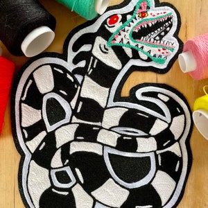 Pode incluir: Um remendo bordado em preto e branco de um monstro de desenho animado com uma boca grande e um olho vermelho. O monstro está em posição enroscada e tem um padrão de listras pretas e brancas.